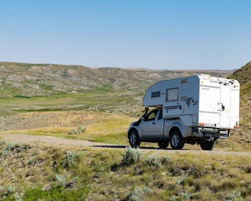 n pick-up Nissan Navara blanc équipé d'une cellule camping-car roulant sur un chemin de terre à travers un paysage vallonné et sec sous un ciel bleu.