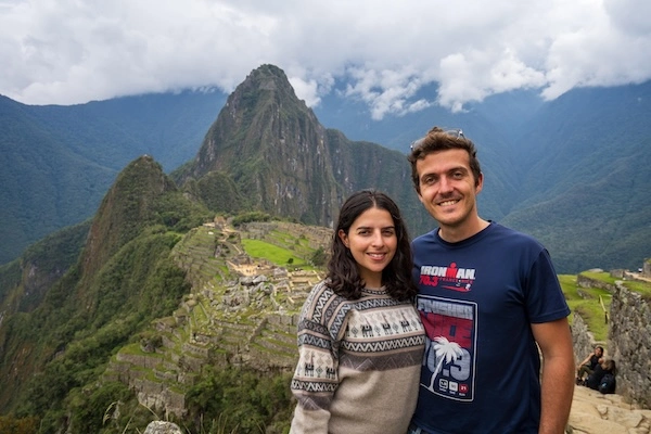 Deux personnes souriantes posant devant le site archéologique du Machu Picchu et les montagnes environnantes sous un ciel nuageux