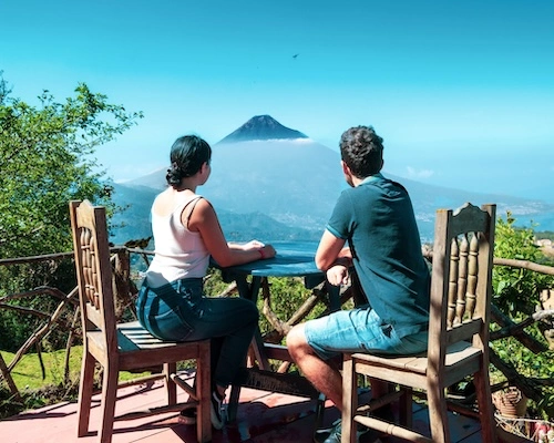 Couple assis à une table en bois sur une terrasse, admirant un volcan au loin dans un paysage tropical
